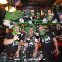 Drei Männer mit grünen Bärten und Hüten sitze an der Theke und halten ein Bier in der Hand
